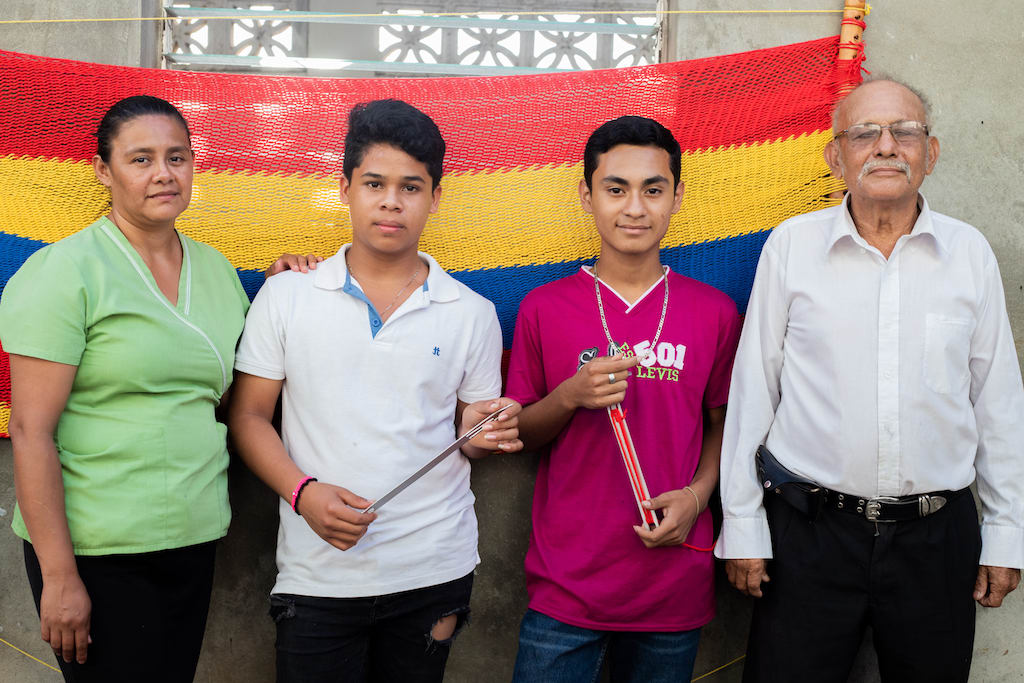 Two teenage boys stand with their tutor and instructor in front of a completed hammock hanging on a wall behind them.
